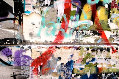 A colorful collage of peeling posters and graffiti on a wall, featuring layers of torn paper and various spray-painted shapes and colors. The artwork blends together, creating a chaotic yet visually engaging scene.