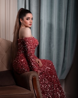 A woman is sitting on a beige upholstered armchair, wearing a sparkling red sequin gown. Her hair is styled in a high ponytail, and she has makeup accentuating her eyes and lips. The background consists of teal curtains and a softly lit room.