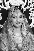 A woman wearing traditional ornate attire with intricate embroidery and jewelry, including a decorative headpiece and a nose ring. The background features elaborate lace or embroidered patterns, contributing to a rich and elegant aesthetic.
