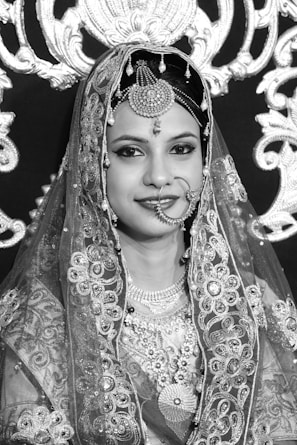 A woman wearing traditional ornate attire with intricate embroidery and jewelry, including a decorative headpiece and a nose ring. The background features elaborate lace or embroidered patterns, contributing to a rich and elegant aesthetic.