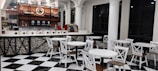 Modern coffee shop interior featuring a bar area with a variety of bottles and a coffee machine on display. The seating area includes several sets of round white tables paired with chairs having white frames and wooden seats. The floor is designed with black and white checkered tiles, and the space is accented with black trim windows and white columns.