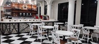 Modern coffee shop interior featuring a bar area with a variety of bottles and a coffee machine on display. The seating area includes several sets of round white tables paired with chairs having white frames and wooden seats. The floor is designed with black and white checkered tiles, and the space is accented with black trim windows and white columns.