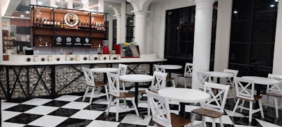 Modern coffee shop interior featuring a bar area with a variety of bottles and a coffee machine on display. The seating area includes several sets of round white tables paired with chairs having white frames and wooden seats. The floor is designed with black and white checkered tiles, and the space is accented with black trim windows and white columns.