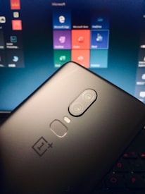 A smartphone with a dual camera is held against the background of a laptop screen displaying various app icons. The phone is a OnePlus model, apparent from its logo. The laptop screen shows a tiled menu with applications like Microsoft Edge, Microsoft Store, and others.