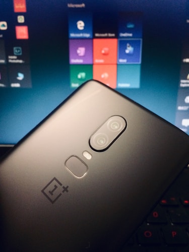 A smartphone with a dual camera is held against the background of a laptop screen displaying various app icons. The phone is a OnePlus model, apparent from its logo. The laptop screen shows a tiled menu with applications like Microsoft Edge, Microsoft Store, and others.