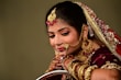A woman is adorned in traditional bridal attire featuring elaborate jewelry, including a nose ring, earrings, and a bejeweled headpiece. She wears a burgundy outfit with gold embellishments and intricate henna designs on her hands.