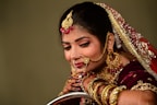 A woman is adorned in traditional bridal attire featuring elaborate jewelry, including a nose ring, earrings, and a bejeweled headpiece. She wears a burgundy outfit with gold embellishments and intricate henna designs on her hands.