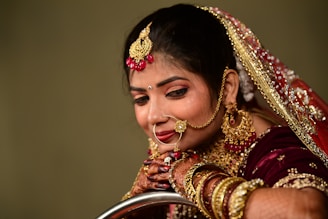 A woman is adorned in traditional bridal attire featuring elaborate jewelry, including a nose ring, earrings, and a bejeweled headpiece. She wears a burgundy outfit with gold embellishments and intricate henna designs on her hands.