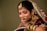 A woman is adorned in traditional bridal attire featuring elaborate jewelry, including a nose ring, earrings, and a bejeweled headpiece. She wears a burgundy outfit with gold embellishments and intricate henna designs on her hands.