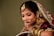 A woman is adorned in traditional bridal attire featuring elaborate jewelry, including a nose ring, earrings, and a bejeweled headpiece. She wears a burgundy outfit with gold embellishments and intricate henna designs on her hands.