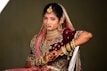 A woman dressed in traditional wedding attire adorned with elaborate jewelry and intricate embroidery. The ensemble includes a richly decorated maroon lehenga with gold and pink embellishments, a matching dupatta draped over her head, and numerous gold bangles on her arm. Her hands are decorated with henna designs, and she wears a large nose ring, necklace, and earrings.