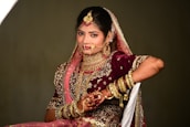 A woman dressed in traditional wedding attire adorned with elaborate jewelry and intricate embroidery. The ensemble includes a richly decorated maroon lehenga with gold and pink embellishments, a matching dupatta draped over her head, and numerous gold bangles on her arm. Her hands are decorated with henna designs, and she wears a large nose ring, necklace, and earrings.