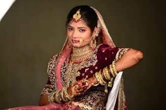 A woman dressed in traditional wedding attire adorned with elaborate jewelry and intricate embroidery. The ensemble includes a richly decorated maroon lehenga with gold and pink embellishments, a matching dupatta draped over her head, and numerous gold bangles on her arm. Her hands are decorated with henna designs, and she wears a large nose ring, necklace, and earrings.