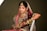 A woman dressed in traditional wedding attire adorned with elaborate jewelry and intricate embroidery. The ensemble includes a richly decorated maroon lehenga with gold and pink embellishments, a matching dupatta draped over her head, and numerous gold bangles on her arm. Her hands are decorated with henna designs, and she wears a large nose ring, necklace, and earrings.