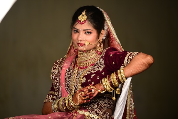 A woman dressed in traditional wedding attire adorned with elaborate jewelry and intricate embroidery. The ensemble includes a richly decorated maroon lehenga with gold and pink embellishments, a matching dupatta draped over her head, and numerous gold bangles on her arm. Her hands are decorated with henna designs, and she wears a large nose ring, necklace, and earrings.