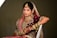 A woman dressed in traditional wedding attire adorned with elaborate jewelry and intricate embroidery. The ensemble includes a richly decorated maroon lehenga with gold and pink embellishments, a matching dupatta draped over her head, and numerous gold bangles on her arm. Her hands are decorated with henna designs, and she wears a large nose ring, necklace, and earrings.