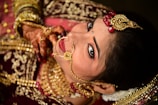 A woman is adorned in intricate gold jewelry including a nose ring and headpiece, featuring red gem accents. Her hands display henna designs, and she wears rich red and gold traditional attire. Her makeup is immaculate, with a focus on the eyes and a small bindi on her forehead.