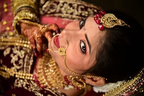 A woman is adorned in intricate gold jewelry including a nose ring and headpiece, featuring red gem accents. Her hands display henna designs, and she wears rich red and gold traditional attire. Her makeup is immaculate, with a focus on the eyes and a small bindi on her forehead.