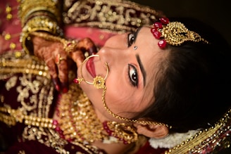 A woman is adorned in intricate gold jewelry including a nose ring and headpiece, featuring red gem accents. Her hands display henna designs, and she wears rich red and gold traditional attire. Her makeup is immaculate, with a focus on the eyes and a small bindi on her forehead.