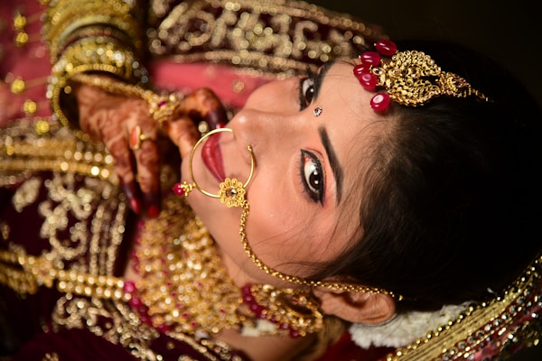 A woman is adorned in intricate gold jewelry including a nose ring and headpiece, featuring red gem accents. Her hands display henna designs, and she wears rich red and gold traditional attire. Her makeup is immaculate, with a focus on the eyes and a small bindi on her forehead.
