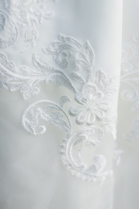 Intricate white embroidery featuring floral and swirl patterns on a slightly translucent fabric. The embroidery is detailed and delicate, creating a sophisticated and elegant texture.