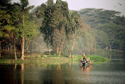 A serene lake surrounded by lush greenery with several trees lining the shore. In the middle of the lake, a small group of people wearing helmets and safety vests navigate a makeshift raft using long poles. Birds can be seen in the sky, adding to the tranquil and natural setting.