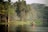 A serene lake surrounded by lush greenery with several trees lining the shore. In the middle of the lake, a small group of people wearing helmets and safety vests navigate a makeshift raft using long poles. Birds can be seen in the sky, adding to the tranquil and natural setting.