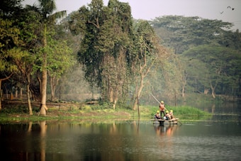 A serene lake surrounded by lush greenery with several trees lining the shore. In the middle of the lake, a small group of people wearing helmets and safety vests navigate a makeshift raft using long poles. Birds can be seen in the sky, adding to the tranquil and natural setting.