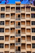 A mid-rise apartment building with a central column of small balconies. The facade is beige with multiple windows, some of which appear to be covered or boarded up. Purple flowering branches are visible in the foreground against a bright blue sky.