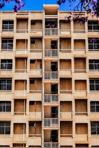 A mid-rise apartment building with a central column of small balconies. The facade is beige with multiple windows, some of which appear to be covered or boarded up. Purple flowering branches are visible in the foreground against a bright blue sky.