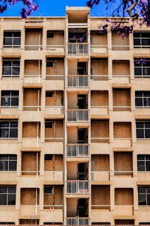 A mid-rise apartment building with a central column of small balconies. The facade is beige with multiple windows, some of which appear to be covered or boarded up. Purple flowering branches are visible in the foreground against a bright blue sky.