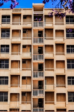 A mid-rise apartment building with a central column of small balconies. The facade is beige with multiple windows, some of which appear to be covered or boarded up. Purple flowering branches are visible in the foreground against a bright blue sky.