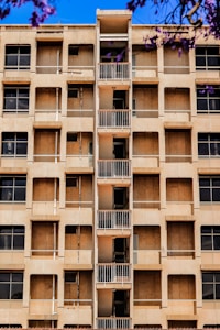 A mid-rise apartment building with a central column of small balconies. The facade is beige with multiple windows, some of which appear to be covered or boarded up. Purple flowering branches are visible in the foreground against a bright blue sky.