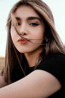 A close-up portrait of a person with long hair flowing across the face, looking directly at the camera. The individual has a distinct makeup style, featuring bold eyebrows and eyeliner with a touch of red. The background is subtle, with soft colors emphasizing the subject's features.