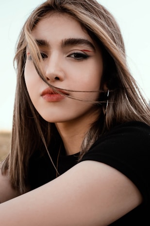 A close-up portrait of a person with long hair flowing across the face, looking directly at the camera. The individual has a distinct makeup style, featuring bold eyebrows and eyeliner with a touch of red. The background is subtle, with soft colors emphasizing the subject's features.