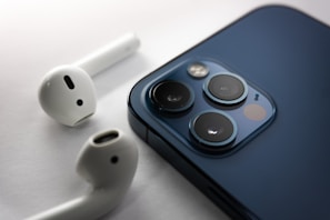A close-up image of a blue smartphone and two white wireless earphones on a white surface. The smartphone's back is visible, showcasing three camera lenses and a flash.