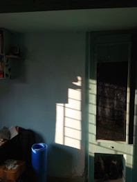 The image captures a sunlit room with shadows cast by a window onto a light blue wall. On the left, there is a shelf with books and a small speaker. Below, a blue yoga mat stands upright next to a small table with a newspaper and boxes. A tall cabinet with glass doors is partially visible, reflecting the light and shadows.