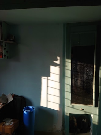 The image captures a sunlit room with shadows cast by a window onto a light blue wall. On the left, there is a shelf with books and a small speaker. Below, a blue yoga mat stands upright next to a small table with a newspaper and boxes. A tall cabinet with glass doors is partially visible, reflecting the light and shadows.