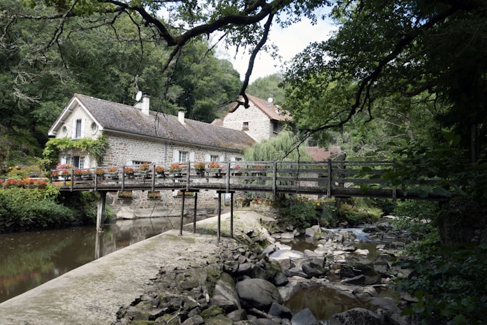 A picturesque stone house surrounded by lush trees and greenery. A wooden bridge adorned with vibrant flowers crosses over a tranquil stream filled with rocks. The setting feels serene and secluded, nestled within a dense forest.