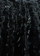 A dark and textured surface with subtle highlights, possibly resembling the look of flowing water or liquid due to its glossy appearance and intricate patterns.