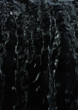 A dark and textured surface with subtle highlights, possibly resembling the look of flowing water or liquid due to its glossy appearance and intricate patterns.