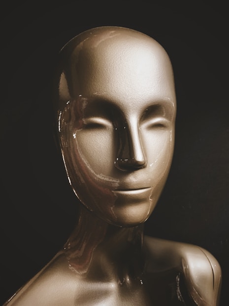 A glossy, metallic mannequin head and upper torso are displayed against a dark background. The mannequin has a smooth surface with abstract, minimal facial features, reflecting light across its contours.