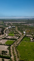 An expansive agricultural landscape with neatly arranged plots of land, bordered by rows of trees. The fields are a mix of green and brown, with some areas left fallow. A few buildings are scattered throughout, providing structure amidst the rural scenery. Dirt roads run parallel to the plots, enabling access across the farmland. A clear blue sky stretches above, enhancing the open, tranquil atmosphere.
