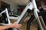 A close-up view of a person interacting with a white electric bicycle, focusing on the battery compartment labeled 'Evelo'. The setting appears to be indoors in a bicycle shop, with several other bicycles visible in the background.