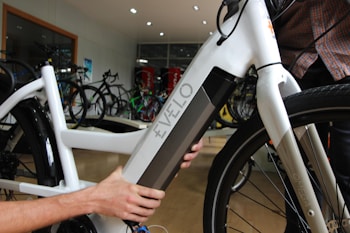 A close-up view of a person interacting with a white electric bicycle, focusing on the battery compartment labeled 'Evelo'. The setting appears to be indoors in a bicycle shop, with several other bicycles visible in the background.