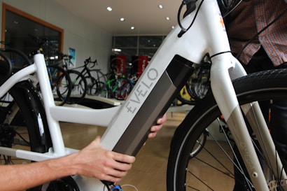 A close-up view of a person interacting with a white electric bicycle, focusing on the battery compartment labeled 'Evelo'. The setting appears to be indoors in a bicycle shop, with several other bicycles visible in the background.