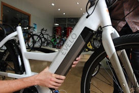 A close-up view of a person interacting with a white electric bicycle, focusing on the battery compartment labeled 'Evelo'. The setting appears to be indoors in a bicycle shop, with several other bicycles visible in the background.