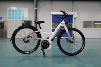 A white electric bicycle stands on a turquoise floor inside a spacious, industrial-like room. The bike features a sleek frame with black tires and a mounted battery. In the background are doors and windows set into light-colored walls, along with a chart or sign on one wall.