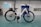 A white electric bicycle stands on a turquoise floor inside a spacious, industrial-like room. The bike features a sleek frame with black tires and a mounted battery. In the background are doors and windows set into light-colored walls, along with a chart or sign on one wall.