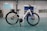 A white electric bicycle stands on a turquoise floor inside a spacious, industrial-like room. The bike features a sleek frame with black tires and a mounted battery. In the background are doors and windows set into light-colored walls, along with a chart or sign on one wall.